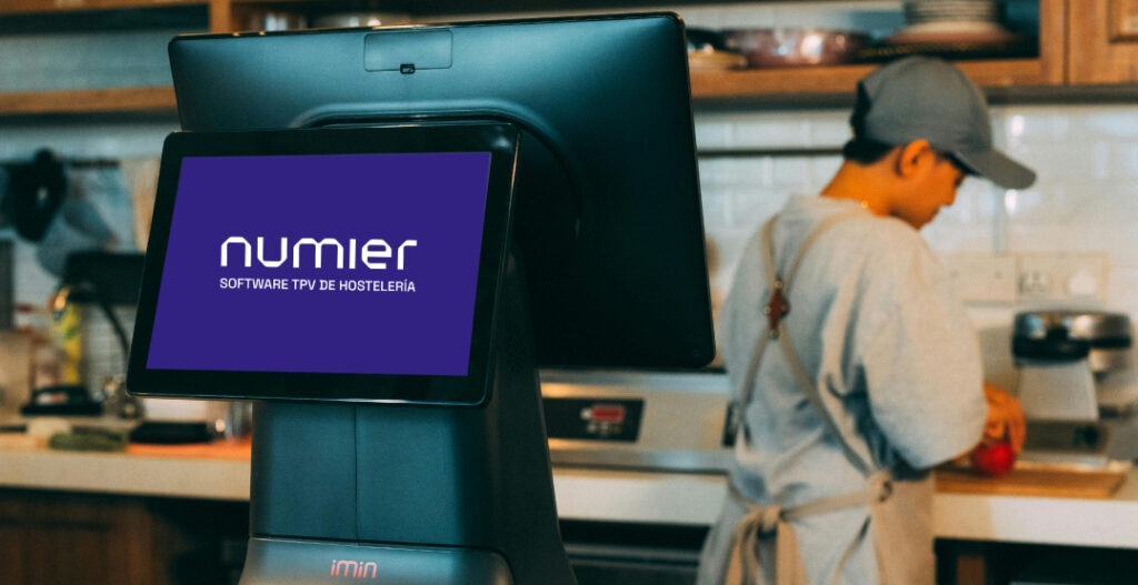Un terminal de Punto de Venta (TPV) iMin moderno de color negro en primer plano, con la pantalla encendida mostrando el logotipo de Numier Software TPV de Hostelería sobre un fondo morado. Al fondo, desenfocado, se ve a un empleado de cocina con delantal y gorra trabajando en una barra de madera con estantes y utensilios de cocina. La imagen evoca un entorno de hostelería profesional.